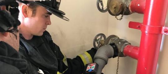 Firefighter attaching equipment to a standpipe system with red pipes and manual control valves.
