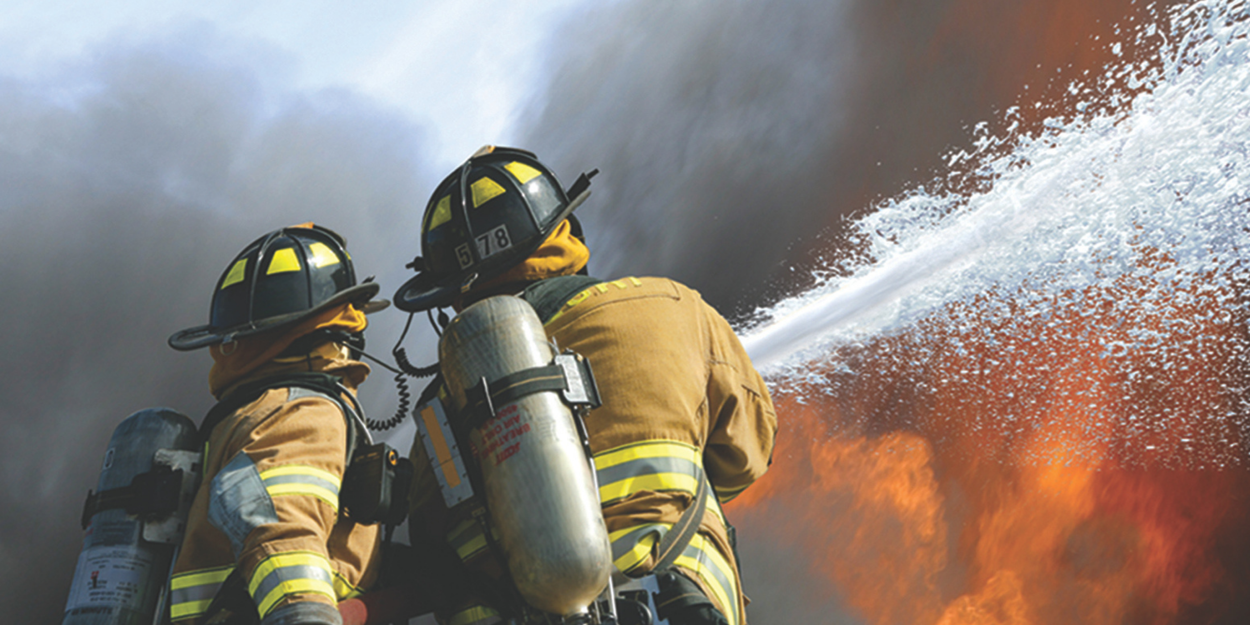Firefighters in protective gear directing a foam and water stream toward an active fire.
